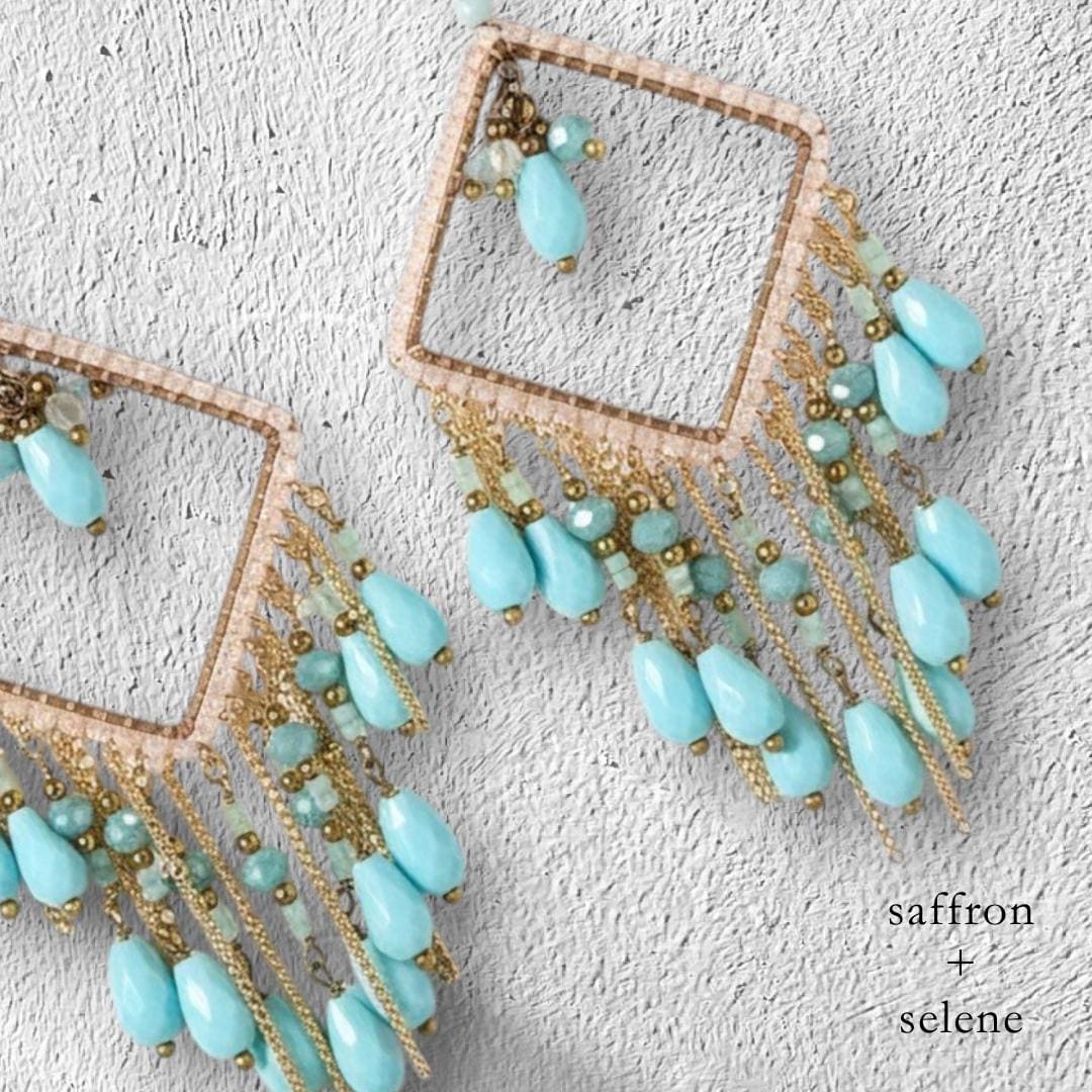 Close-up of two turquoise and gold-toned earrings. Each earring features a square frame with dangling turquoise teardrop beads and gold chain accents. The text "saffron + selene" is visible.