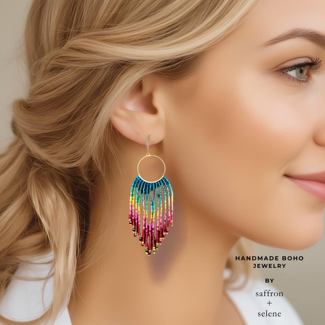 Gold-tone hoop earrings with a colorful beaded fringe. The fringe ombretransitions from teal to yellow, pink, and burgundy. The earrings are a vibrant boho accessory gift for her.